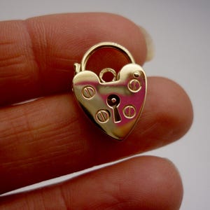 May include: A gold heart-shaped padlock charm with a keyhole and decorative details. The charm is shown on a finger.