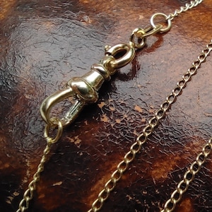 May include: A gold chain with a decorative clasp. The chain is made of small, linked circles. The clasp is a hook with a decorative loop.