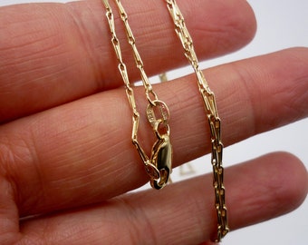 May include: A delicate gold necklace with a rectangular link chain. The necklace is shown against a light background. The clasp is visible, and the chain is held in a hand.