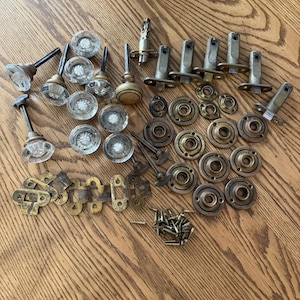 May include: A collection of antique door hardware, including glass and brass doorknobs, metal backplates, hinges, and screws. The hardware is arranged on a wooden surface, showcasing various styles and finishes.