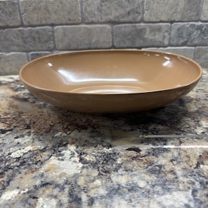 May include: A smooth, solid tan-colored shallow bowl with a wide, rounded rim. The bowl features a glossy finish that reflects light across its curved interior surface, presenting a simple, circular design with no additional patterns or markings.