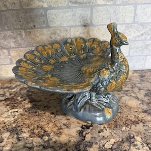 May include: A ceramic peacock-shaped dish featuring a glossy finish with a blend of slate blue and golden yellow glaze. The bird's tail feathers fan out to form the bowl, while the head and neck are upright, resting on a sturdy, rounded base.