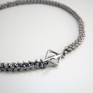 May include: A silver-toned necklace with a woven chain design. The necklace features a geometric clasp. The chain is made of small, interconnected metal rings, creating a textured appearance. The jewelry is displayed on a white surface.