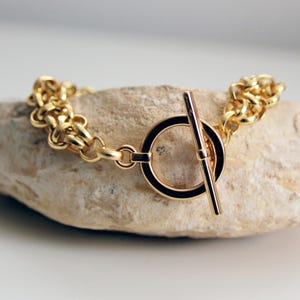 May include: A gold chain bracelet with a toggle clasp. The bracelet is on a white rock.