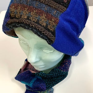 Puede incluir: Un gorro de punto azul y marrón con un calientaorejas de forro polar azul. El gorro se combina con una bufanda de punto azul y verde y guantes sin dedos a juego.
