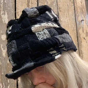 Op de afbeelding: Een zwarte en witte patchwork gebreide muts met een brede rand. De muts is gemaakt van verschillende texturen en patronen van gebreid stof.