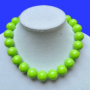 May include: A vibrant lime green beaded necklace. The necklace features large, glossy, round beads strung together. The beads are displayed on a white mannequin bust against a blue background. This statement necklace is a bold accessory.