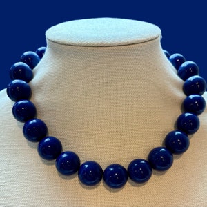 May include: A necklace made of large, round, dark blue beads. The necklace is displayed on a white fabric mannequin.