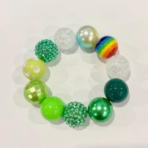 May include: A collection of colorful beads arranged in a circle. The beads vary in size, color, and texture, including green, white, yellow, and a rainbow-striped bead. Some beads have a glittery or textured surface.