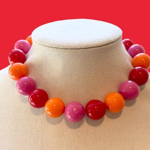 May include: A colorful beaded necklace featuring large, round beads in shades of red, pink, and orange. The necklace is displayed on a white bust against a red background. The beads appear to be approximately 1 inch in diameter.