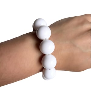 May include: A white beaded bracelet with large, round beads. The bracelet is worn on a wrist and is against a white background. The beads appear to be made of a glossy material.