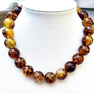 May include: A chunky necklace featuring large, round beads in shades of amber and brown. The beads have a textured, crackled appearance, creating a unique and eye-catching design. The necklace is displayed on a white bust, showcasing its bold and elegant style.