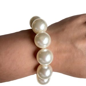 Może przedstawiać: Kremowa bransoletka z pereł. Bransoletka składa się z dużych, okrągłych pereł nanizanych na sznurek. Perły mają błyszczące wykończenie i są prezentowane na jasnym tle. Klasyczny element biżuterii.