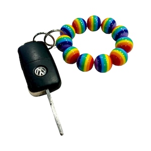 Puede incluir: Un llavero de cuentas de colores arcoíris con una llave de coche negra adjunta. El llavero está hecho de cuentas redondas con rayas alternas de rojo, naranja, amarillo, verde, azul, índigo y violeta.
