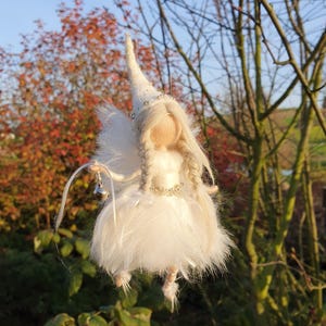 Könnte beinhalten: Eine weiße Feenpuppe mit einem langen weißen Kleid, weißen Flügeln und einer weißen spitzen Haube. Die Puppe hat blonde Haare und eine Perlenkrone. Die Puppe hängt an einem Ast vor einem verschwommenen Hintergrund aus Bäumen und Himmel.