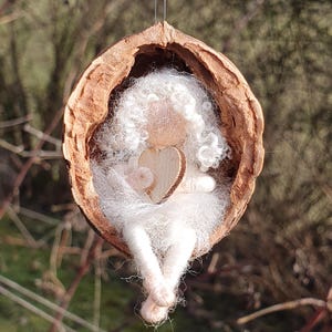Op de afbeelding: Een witte vilten feepop met een houten hart, zittend in een uitgeholde walnootschelp.