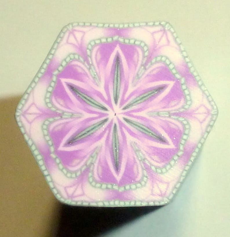 May include: A hexagonal purple and white polymer clay tile with a detailed floral design. The center of the tile features a six-pointed star with a white center.