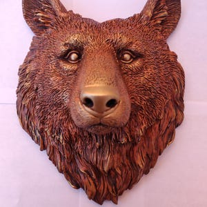 Può includere: Una scultura da parete raffigurante una testa di orso con una finitura testurizzata color bronzo. L'oggetto presenta dettagliati motivi del pelo, orecchie a punta e una zona del naso e degli occhi liscia, che riproduce i tratti di un orso.