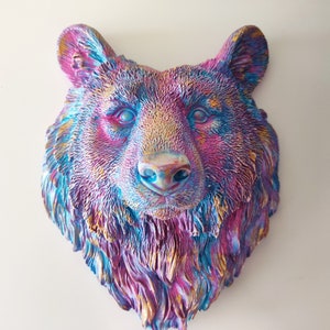 May include: A wall-mounted bear head sculpture featuring a textured fur finish. The surface is painted in a vibrant, iridescent colour palette of metallic pink, purple, blue, and gold, creating a multi-dimensional effect across the bear's face.