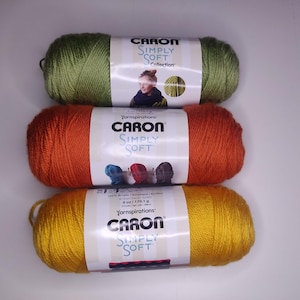 Può includere: Tre gomitoli di filato Caron Simply Soft impilati verticalmente. Il gomitolo superiore è verde oliva, quello centrale è arancione bruciato e quello inferiore è giallo oro. Ogni gomitolo presenta un'etichetta di carta bianca con un peso di 170,1 g.
