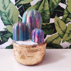 May include: A trio of colourful, hand-painted concrete cacti in a gold pot. The cacti are painted in shades of blue, purple and pink with a glittery finish.