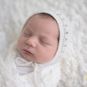 May include: A newborn baby sleeping peacefully in a white crocheted blanket and hat. The baby's face is visible, with closed eyes and a soft expression.