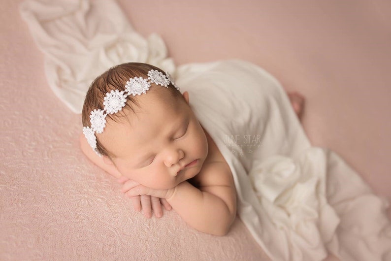 May include: A newborn baby girl sleeping on a pink textured background, wearing a white crocheted flower headband. The baby is wrapped in a white blanket.