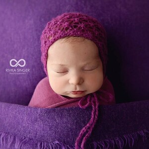 May include: A newborn baby wrapped in a purple blanket and wearing a knitted magenta bonnet. The baby is nestled in a deep purple fabric, with the photographer's logo in the upper left corner.