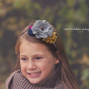May include: A young girl with long brown hair is wearing a headband with a gray, pink, and blue fabric flower. The headband is decorated with a small, sparkly red jewel. The girl is smiling and looking to the right. The background is blurred and out of focus.