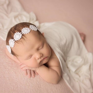 May include: A newborn baby girl sleeping on a pink textured background, wearing a white crocheted flower headband. The baby is wrapped in a white blanket.