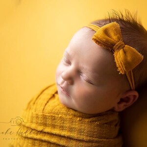 May include: A newborn baby wrapped in a mustard yellow blanket, wearing a matching headband with a bow. The baby is sleeping peacefully against a yellow backdrop. The image is a studio portrait.