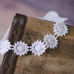 May include: White lace trim with a scalloped edge and a repeating flower design. The trim is on a rustic wooden surface.