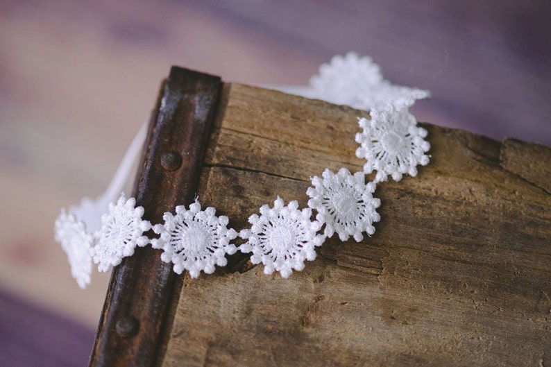 May include: White lace trim with a scalloped edge and a repeating flower design. The trim is on a rustic wooden surface.