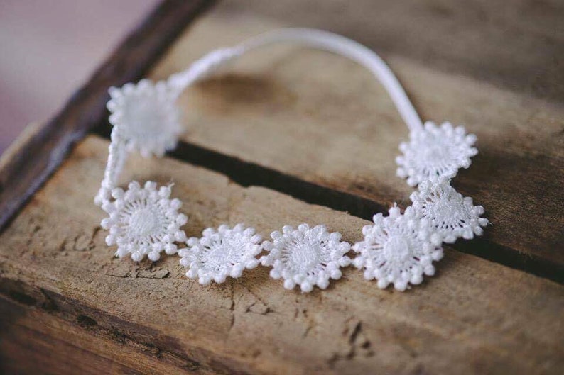 May include: White lace headband with a delicate floral design. The headband is made of white lace and features a series of small, white flowers.