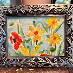 May include: A rectangular picture frame with a metallic silver finish and an ornate leaf and scrollwork border. Inside the frame is a watercolor painting depicting yellow flowers and a single red poppy against a light blue background.