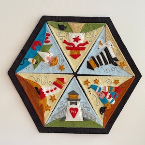 May include: A felt hexagon wall hanging with six triangular sections, each featuring a different whimsical design. The designs include a lobster, a lighthouse with "PEI" on it, and a stack of buoys. The border is black.