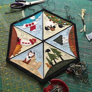 May include: A handmade, hexagonal felt wall hanging with six triangular scenes. Each scene depicts a whimsical landscape with felted figures and details. The piece is surrounded by a black border. Scissors, thread, and pins are nearby.