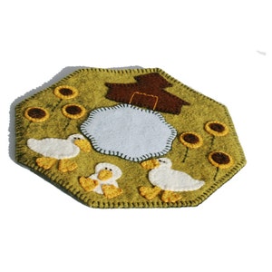 Puede incluir: Una aplicación de fieltro de un estanque con tres patos y una pequeña casa. El estanque está rodeado de girasoles sobre un fondo verde.