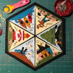 May include: A hexagonal felt wall hanging featuring six different scenes. The scenes include a lighthouse, a house, a tree, a fox, a boat with oars, and a group of seagulls. The scenes are all surrounded by a black border and are stitched with a variety of colours.