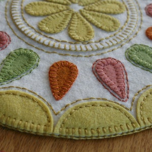 Op de afbeelding: Een witte viltcirkel met een gestikt ontwerp van een bloem in het midden en traanvormige vormen rond de rand. De traanvormige vormen zijn in tinten groen, oranje en roze.