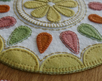 May include: A white felt circle with a stitched design of a flower in the center and teardrop shapes around the edge. The teardrop shapes are in shades of green, orange, and pink.