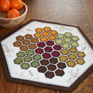 May include: A white felt table runner with a brown felt border. The center of the runner features a honeycomb design with hexagons in shades of brown, orange, green, and burgundy.