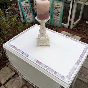 May include: White drop leaf table with a purple and white mosaic tile border. The table has a white candle holder with a pink candle on top.