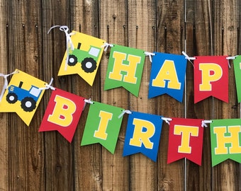 May include: A two-row Happy Birthday banner featuring colorful pennants in red, blue, green, and yellow. The banner includes illustrations of blue, green, and red tractors on the end pieces, all strung together with white ribbon.