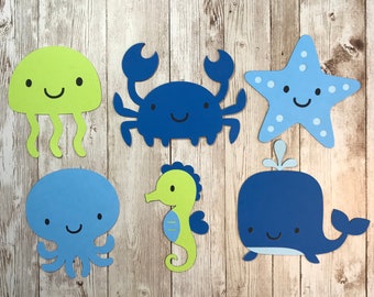 May include: A set of six paper cutouts featuring sea creatures including a lime green jellyfish, a dark blue crab, a light blue starfish, a light blue octopus, a lime green seahorse, and a dark blue whale with a water spout.
