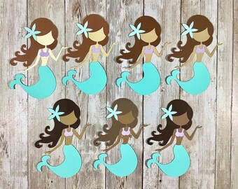May include: Seven paper mermaid cutouts arranged in two rows. Each mermaid features brown hair with a light blue flower, a light purple bikini top, and a bright turquoise tail. The figures are flat, die-cut shapes with simple facial features.