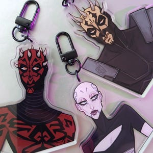 Pode incluir: Três chaveiros com ilustrações de personagens do universo Star Wars. Os chaveiros são feitos de acrílico transparente e têm um anel de metal preto. Os personagens são Darth Maul, Asajj Ventress e o General Grievous.