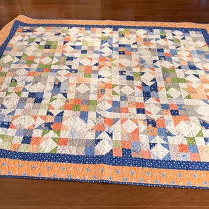 May include: A square patchwork quilt featuring a repeating geometric star pattern in shades of blue, orange, green, and white. The quilt is finished with a dark blue inner border and a wide, solid orange outer border with a floral print.
