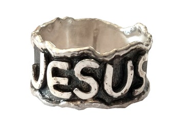 May include: A silver ring with the word "JESUS" embossed on the band. The ring has a textured, rough surface.