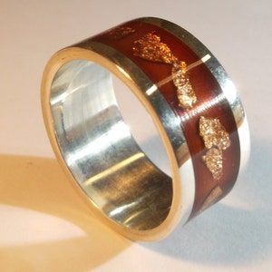May include: A silver band ring with a brown resin inlay featuring gold flakes. The resin is translucent, allowing the gold flakes to be seen through the brown.